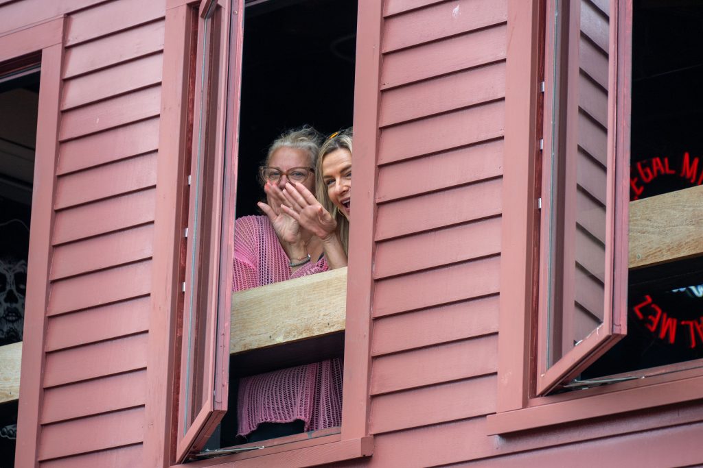 Zwei Frauen winken aus einem geöffneten Fenster einer rosa Holzfassade.